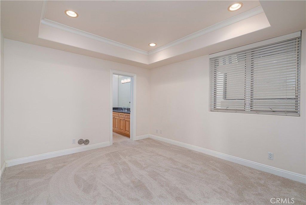 This spacious, neutral-toned bedroom features plush carpeting and a tray ceiling with recessed lighting. A large window with horizontal blinds allows natural light to fill the room, while an open doorway leads into an adjacent bathroom with wooden cabinetry. The room offers a clean, blank canvas aesthetic suitable for a guest bedroom or secondary living space.