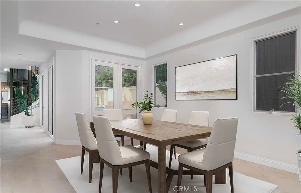 This bright and airy dining room features a light wood table surrounded by six modern, cream-colored upholstered chairs. The space is anchored by a neutral area rug on light-toned flooring, with a large abstract painting serving as a focal point on the wall. Large glass doors provide natural light and a view of the outdoor greenery, creating a clean and inviting atmosphere.