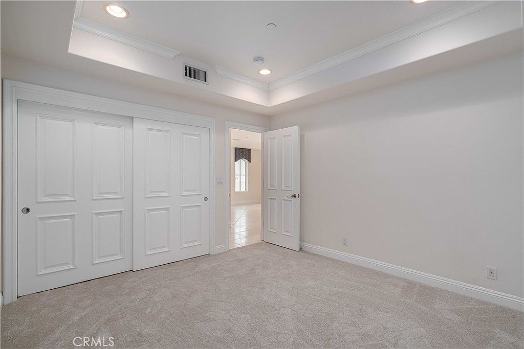 This interior view shows a neutral-toned bedroom featuring light-colored carpeting and white walls. A large set of sliding closet doors occupies the left wall, while an open doorway leads to a bright, adjacent room. The room includes a tray ceiling with recessed lighting, creating a clean and spacious atmosphere.