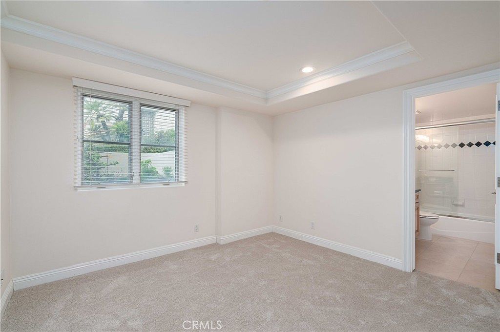 This bright, neutral-toned bedroom features plush carpeting and a tray ceiling that adds architectural interest to the space. A large window with white blinds allows for natural light, while an open doorway leads directly into an attached bathroom with a glass-enclosed shower. The room offers a clean, blank-slate aesthetic suitable for a guest bedroom or home office.