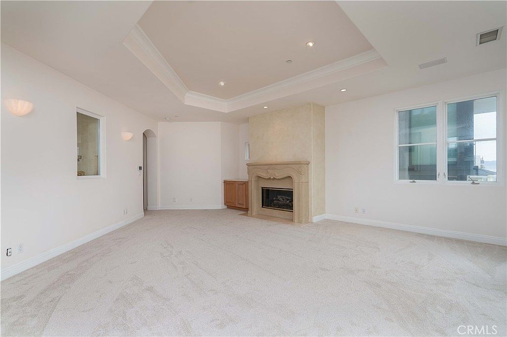 This spacious living room features neutral carpeting and light-colored walls, creating a bright and airy atmosphere. A prominent fireplace with a decorative stone surround serves as the focal point, complemented by a recessed tray ceiling and wall-mounted sconces. Large windows on the right allow for ample natural light, while an arched doorway leads to an adjacent area.