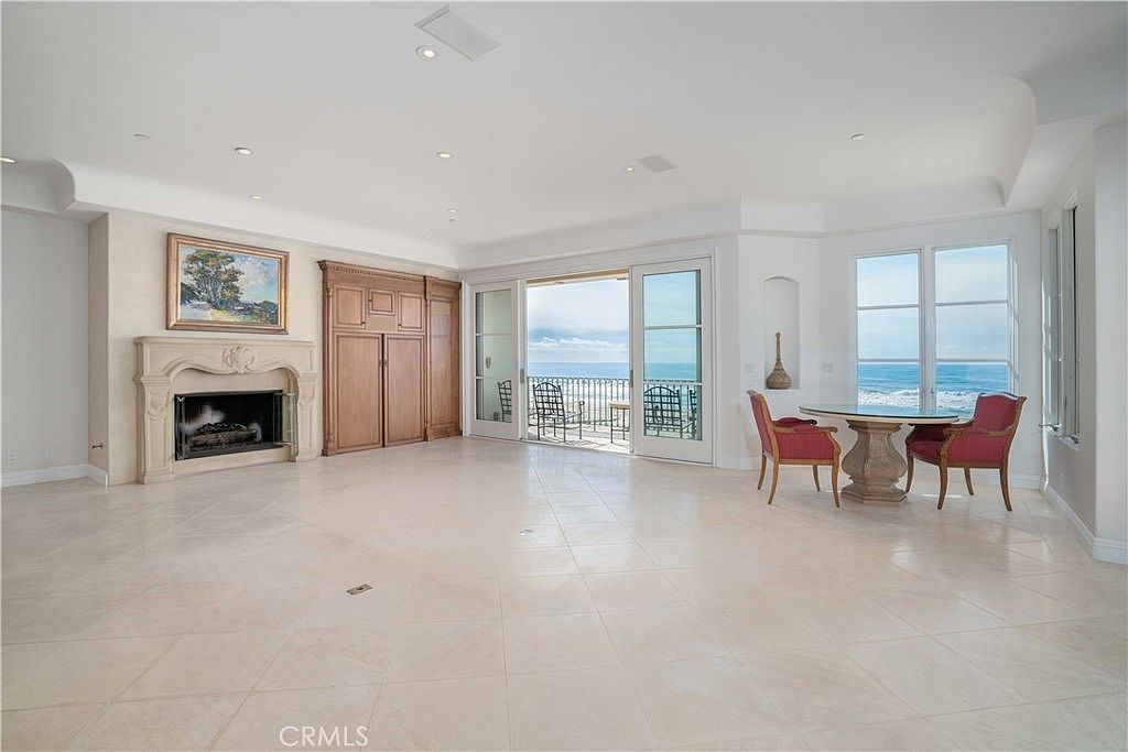 This spacious living room features light-colored tile flooring and a prominent stone fireplace with a decorative mantel. A set of large sliding glass doors opens onto a balcony, providing an expansive, unobstructed view of the ocean. To the right, a small dining area with two red chairs is positioned to take advantage of the coastal scenery.