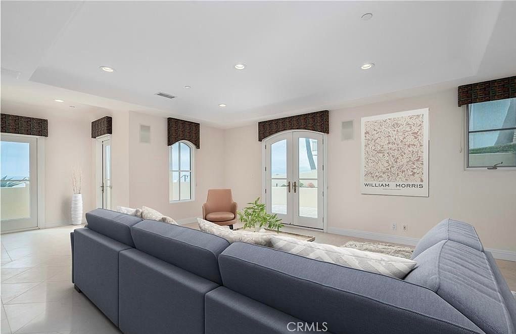 This spacious living room features a large, neutral-toned sectional sofa facing a William Morris art print on the wall. The room is brightened by recessed lighting and multiple sets of French doors that provide access to an outdoor balcony with ocean views. The space is finished with light-colored tile flooring and valanced window treatments, creating a clean and airy atmosphere.