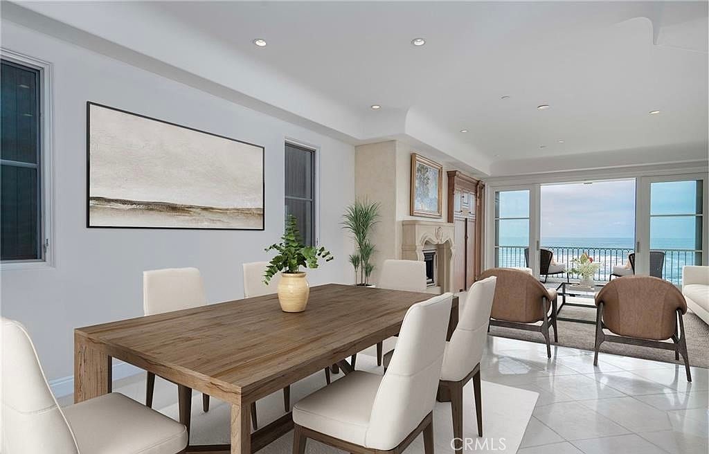 This elegant dining area features a large light wood table surrounded by modern, cream-colored upholstered chairs. The space is bright and airy, with a neutral color palette and large glass doors that open to a balcony with expansive ocean views. A decorative stone fireplace surround adds a touch of warmth to the room, while recessed lighting and a large piece of abstract wall art complete the sophisticated aesthetic.