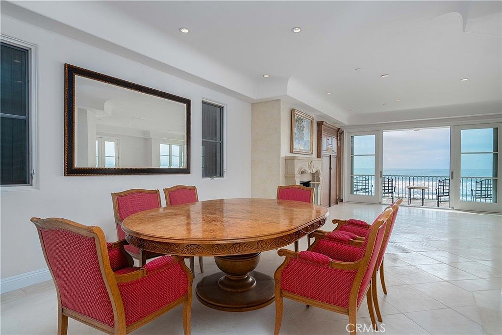 This elegant dining room features a large, round wooden table surrounded by six chairs with red patterned upholstery. A prominent rectangular mirror hangs on the wall, reflecting the space, while a stone fireplace with a decorative mantel adds a classic touch to the room. Large glass doors open to a balcony, providing an expansive view of the ocean and filling the room with natural light.