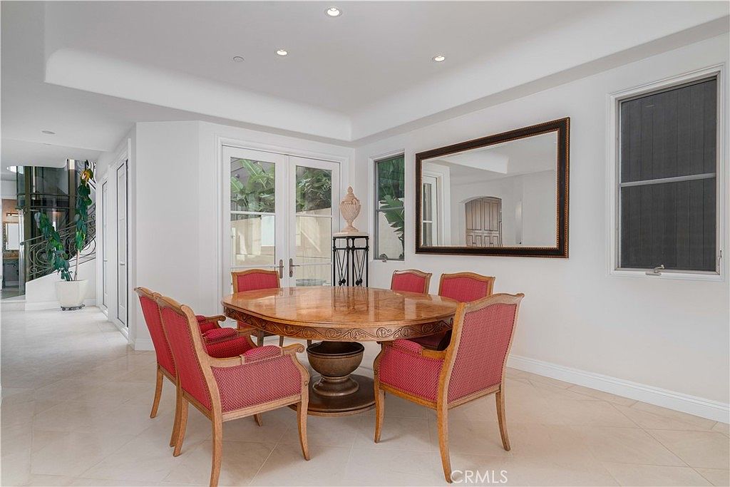 This formal dining room features a large, round wooden table surrounded by six chairs upholstered in a vibrant red fabric. The space is bright and airy, with light-colored tile flooring, recessed ceiling lighting, and a large rectangular mirror mounted on the wall. French doors provide natural light and a view of the outdoor area, creating an elegant and inviting atmosphere.