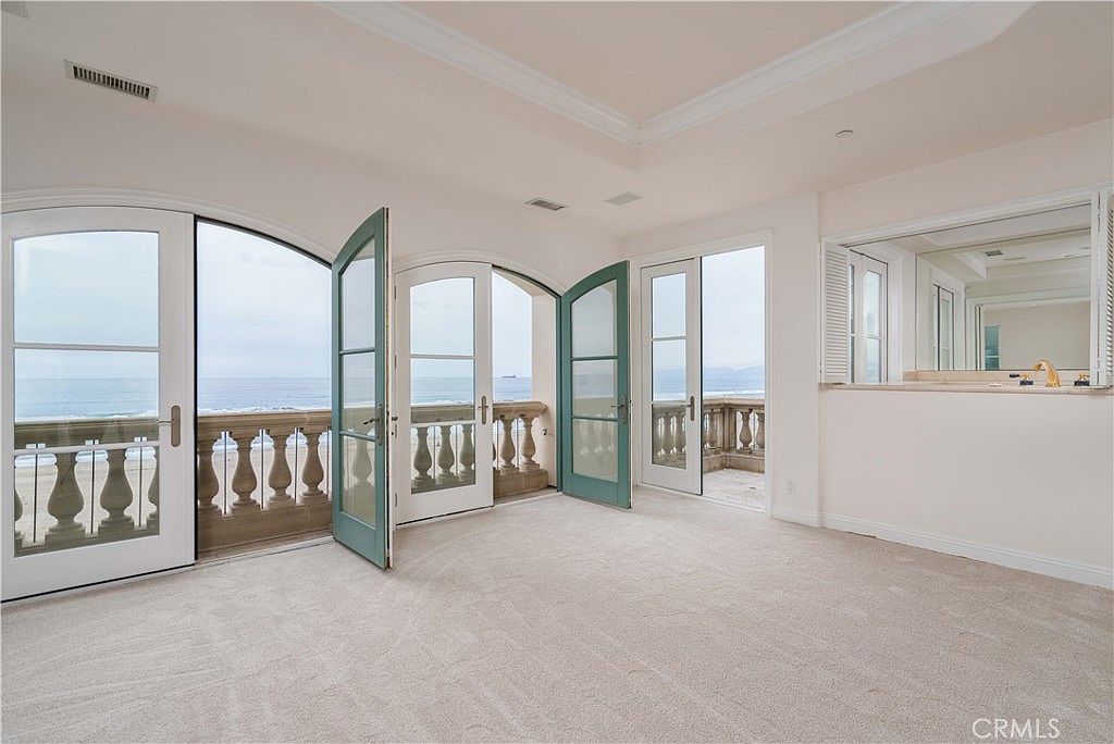 This bright and airy primary bedroom features neutral carpeting and light-colored walls, creating a serene atmosphere. The room is defined by multiple sets of French doors that open onto a private balcony, offering expansive, unobstructed views of the ocean. To the right, a built-in vanity area with a mirror and sink provides a convenient transition space, likely leading to an en-suite bathroom.