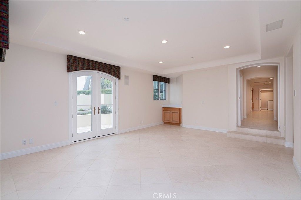 This bright, neutral-toned living room features light-colored tile flooring and recessed lighting throughout the ceiling. A set of double French doors with arched tops provides access to the outdoors, while a small built-in wooden cabinet sits beneath a window on the adjacent wall. The space leads into a hallway, creating an open and airy transition between rooms.