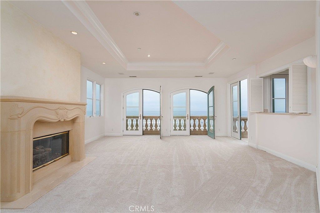 This spacious living room features neutral carpeting and a prominent, ornate stone fireplace surround on the left wall. The room is brightened by multiple sets of French doors that open onto a balcony, providing a clear view of the ocean beyond. The ceiling includes a recessed tray design, adding architectural depth to the airy, light-filled space.