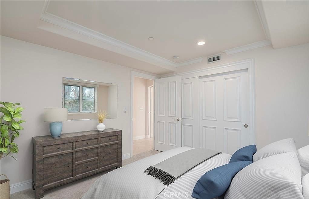 This bright and neutral bedroom features a large bed with blue accent pillows and a textured grey throw blanket. A wooden dresser sits against the wall beneath a rectangular mirror, while a doorway leads to an adjacent hallway. The room is finished with white trim, recessed lighting, and a set of white paneled closet doors.