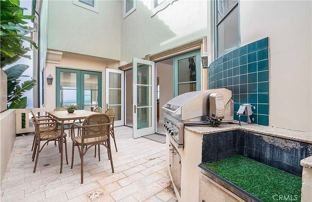 This outdoor patio space features light-colored stone pavers and a built-in stainless steel grill station with a teal tiled backsplash. A dining set with woven chairs sits adjacent to the grill, while multiple sets of glass-paned doors provide seamless access to the interior living areas. The space is framed by light-colored walls and lush greenery, creating a private and inviting atmosphere for al fresco dining.