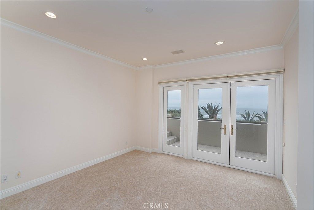 This bright, neutral-toned bedroom features plush carpeting and recessed ceiling lighting. A set of double glass doors provides access to an outdoor balcony, offering a clear view of the ocean and coastal landscaping. The room is minimalist in design, emphasizing the natural light and the scenic exterior connection.