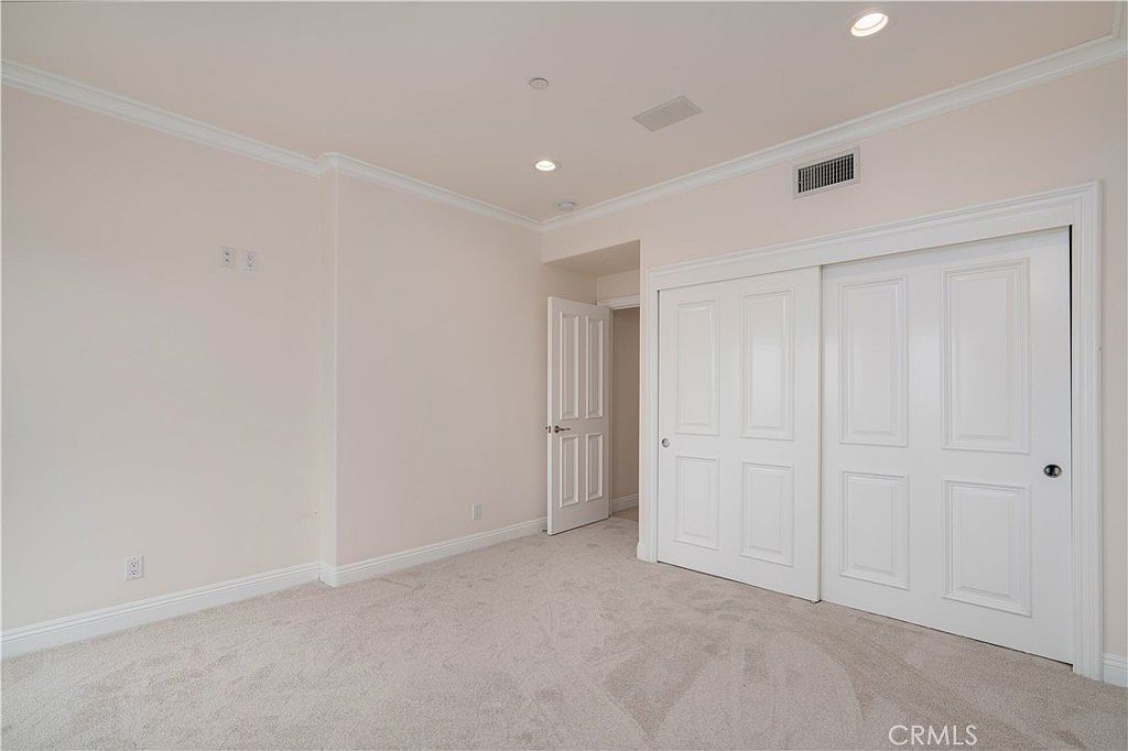 This image shows a neutral-toned, unfurnished bedroom featuring light-colored walls and plush carpeting. A set of white, multi-panel sliding closet doors occupies the right wall, while an open doorway on the left leads to an adjacent space. The room is illuminated by recessed ceiling lights, creating a clean and versatile canvas for potential residents.