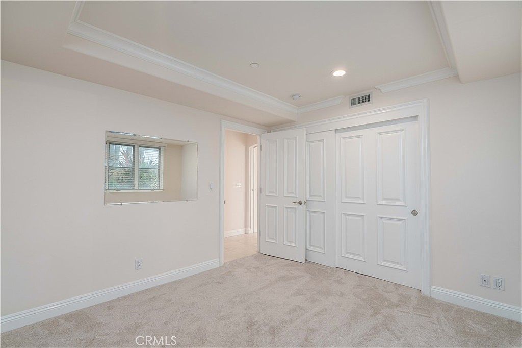 This bright, neutral-toned bedroom features plush wall-to-wall carpeting and a recessed tray ceiling that adds architectural interest. A large, rectangular mirror is mounted on the wall, reflecting light and creating a sense of depth, while a set of double doors leads to a closet. The room is accessed through an open doorway, providing a clear view into the adjacent hallway.