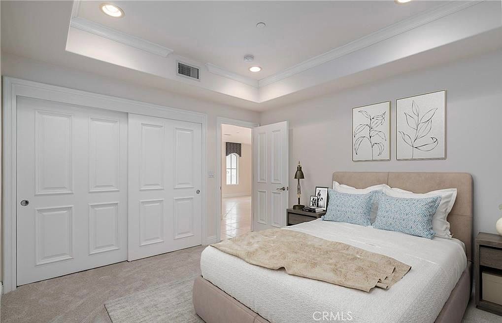 This well-lit bedroom features a neutral color palette with soft gray walls and plush carpeting. The room is furnished with a bed adorned with patterned blue pillows and a light-colored throw, flanked by two nightstands. A tray ceiling adds architectural interest, while a doorway leads to an adjacent room, and a large sliding-door closet provides ample storage.