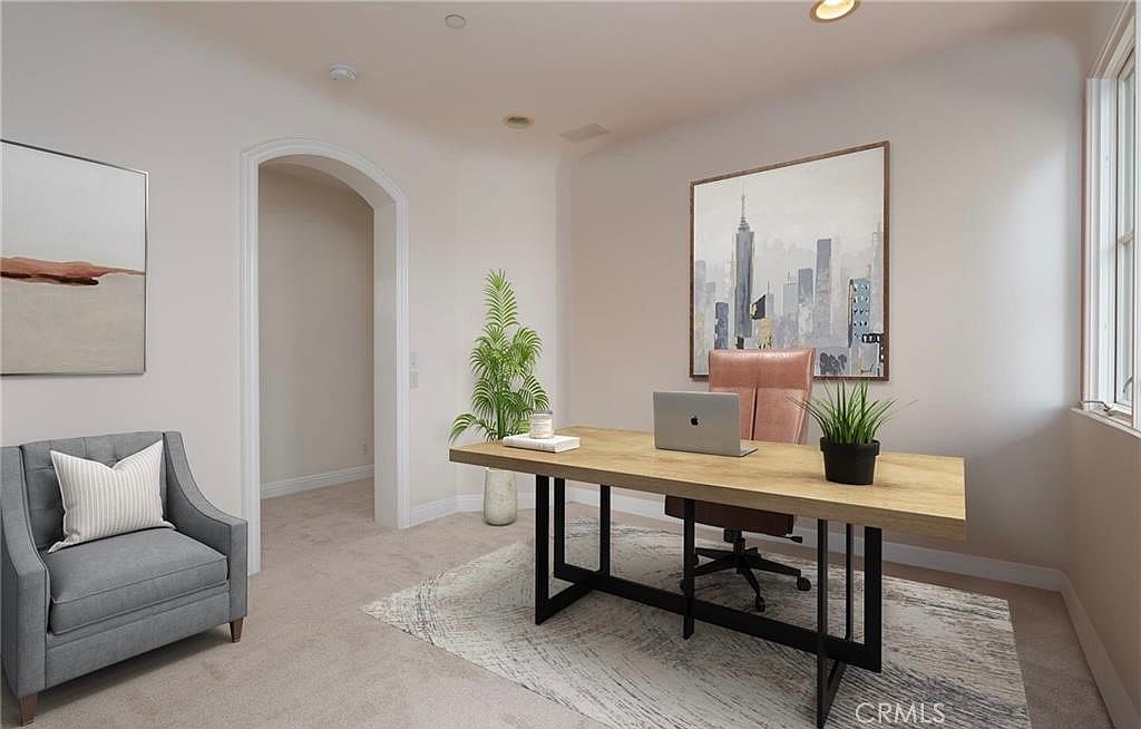 This home office features a light wood desk with a modern black metal frame, paired with a pink upholstered office chair. A gray armchair with a white throw pillow sits to the left, while a potted plant adds a touch of greenery near an arched doorway. The room is decorated with a large cityscape wall art piece and a patterned area rug, creating a professional yet inviting workspace.