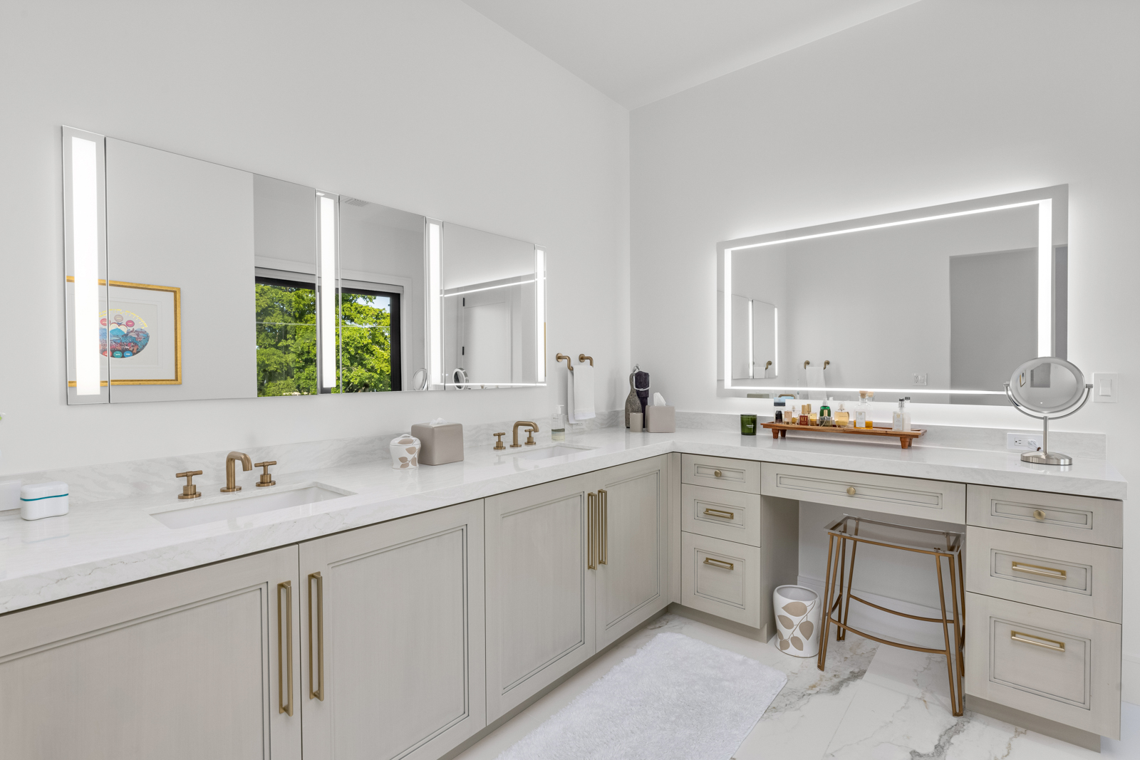 This is a bright and modern primary bathroom featuring a double vanity with a white countertop and light gray cabinetry. The room is illuminated by integrated lighting in the mirrors, and the fixtures are a brushed gold color. A makeup vanity with a stool is also present, adding to the luxurious feel of the space.