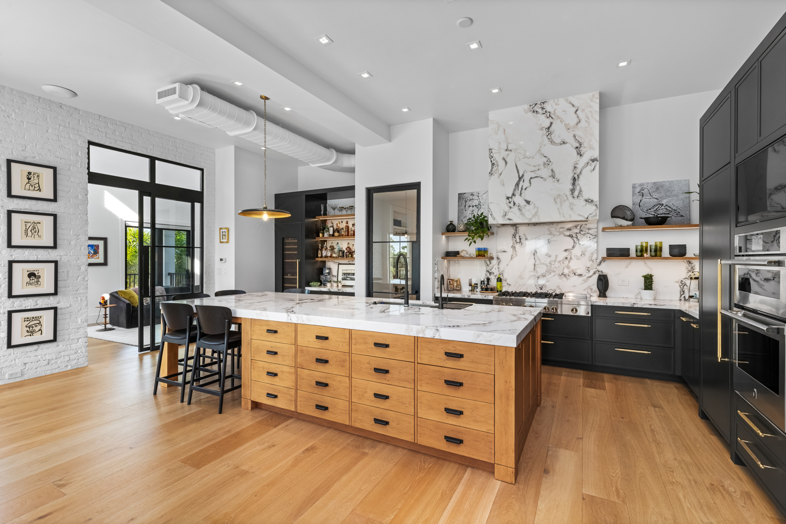 This is a modern, high-end kitchen featuring a large island with a marble countertop and wooden drawers. The kitchen also includes black cabinetry with gold hardware, stainless steel appliances, and a marble backsplash. The open layout and natural light create a bright and inviting space.