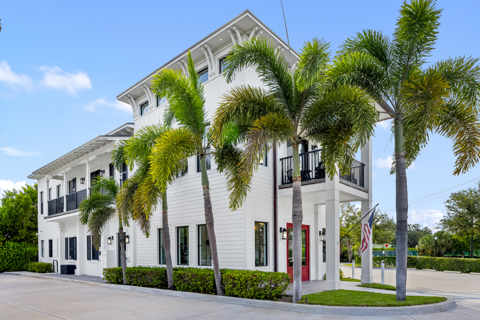 This is a front view of a two-story white building with a red front door and black framed windows. Palm trees line the front of the building, adding a tropical feel. The building has balconies with black railings and a well-maintained lawn, creating a welcoming and upscale impression.