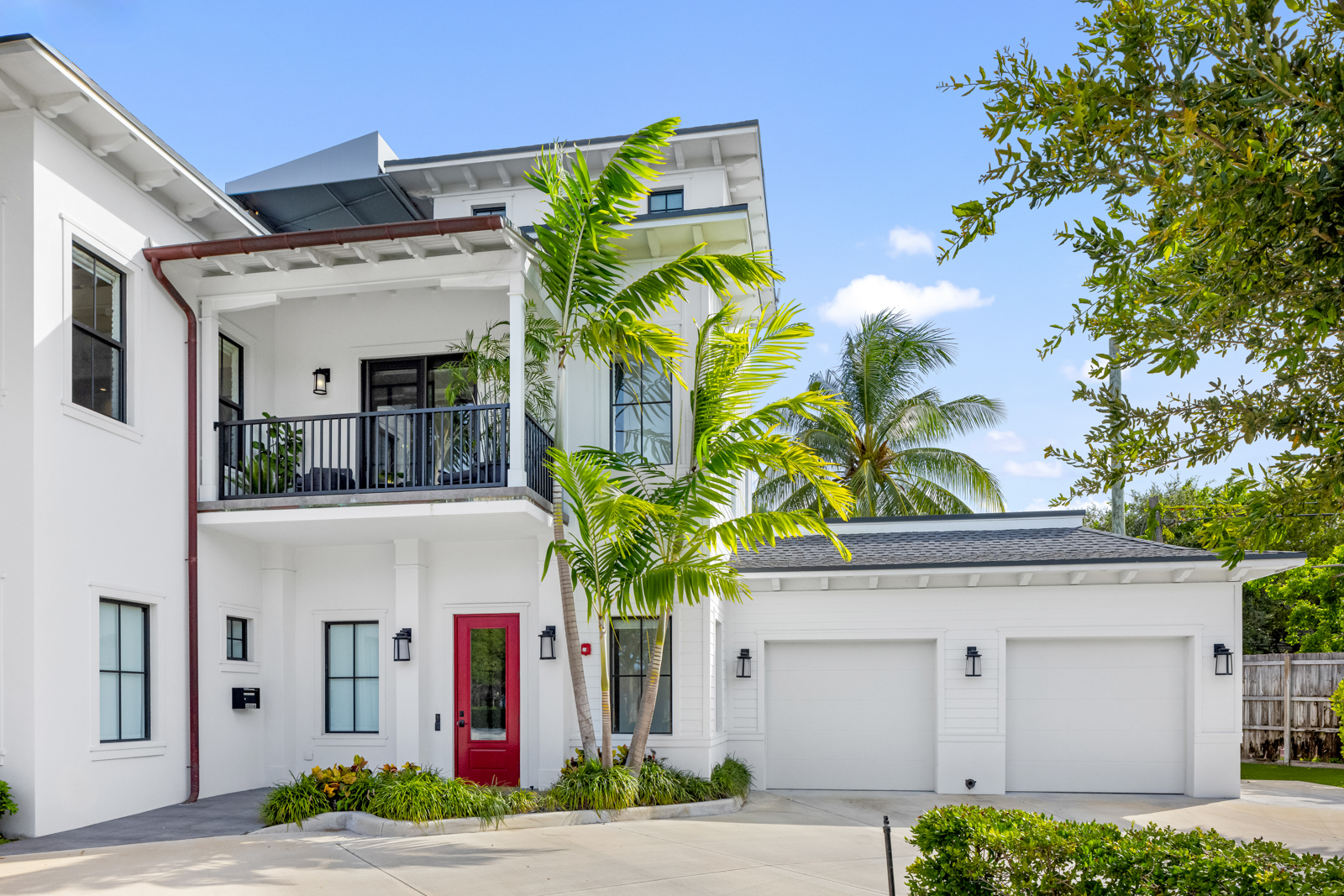 This is a front exterior view of a modern two-story home with a clean, white facade. The house features a red front door, a second-story balcony with black railings, and a two-car garage on the right side. Palm trees and manicured landscaping add to the property's curb appeal, creating a welcoming and upscale impression.