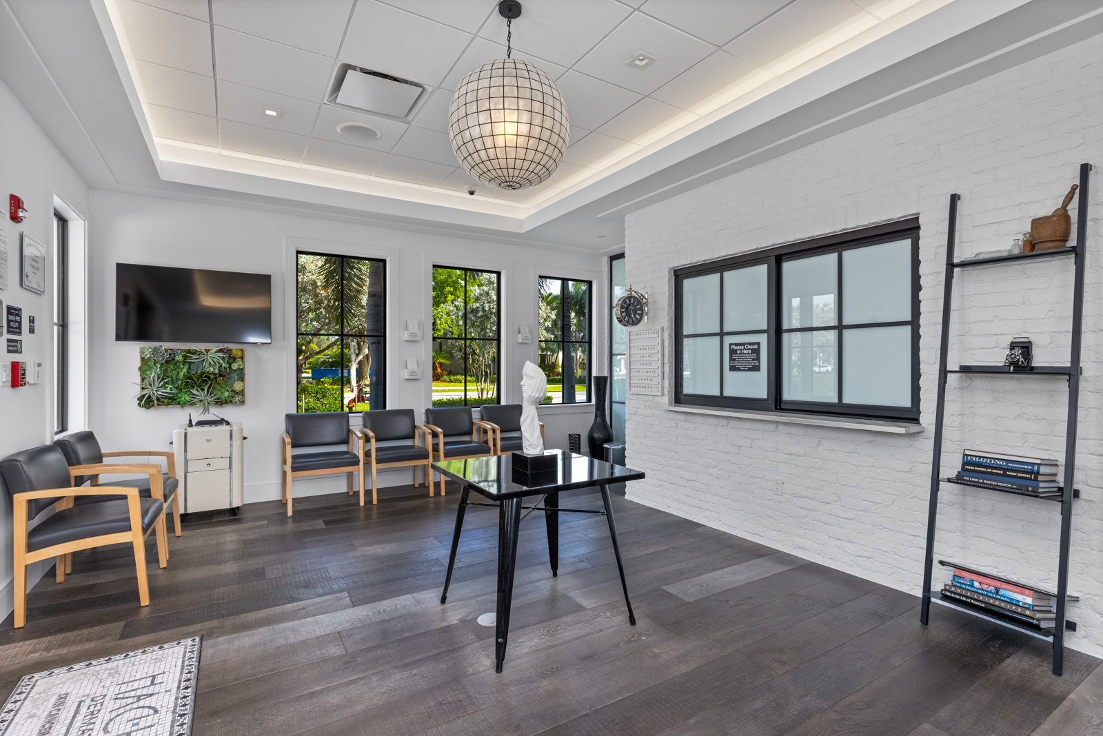 This is a well-lit office or waiting area featuring modern design elements. The room includes comfortable seating, a sleek black table, and a decorative shelving unit. Large windows provide natural light, and the overall aesthetic is clean and professional, suitable for a high-end real estate listing.