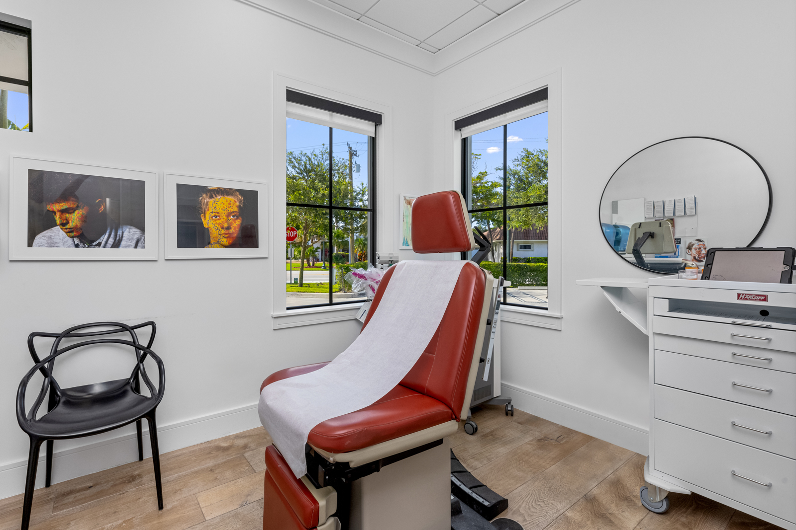 This is an interior shot of a doctor's office or examination room. The room features a red examination chair with a white paper cover, positioned in front of two windows that offer a view of the outside. A black chair sits to the left, and a white cabinet with a round mirror on top is on the right, creating a clean and professional atmosphere.