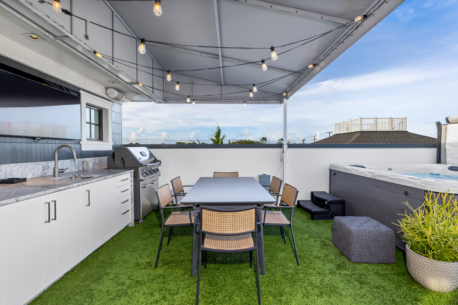 This is a stylish rooftop patio featuring an outdoor kitchen with white cabinets, a stainless steel grill, and a sink. A dining table with six chairs sits on artificial turf, creating a cozy outdoor dining area. A hot tub is also present, suggesting a luxurious and relaxing outdoor space with string lights overhead.