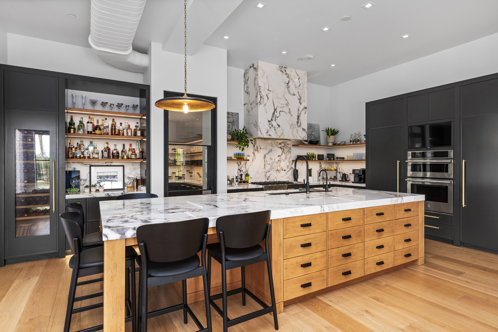 This is a modern kitchen featuring dark cabinetry and a large island with a marble countertop and light wood drawers. The kitchen includes a wine fridge, built-in ovens, and a marble range hood. The space is well-lit with recessed lighting and a pendant light over the island, creating a luxurious and functional cooking area.
