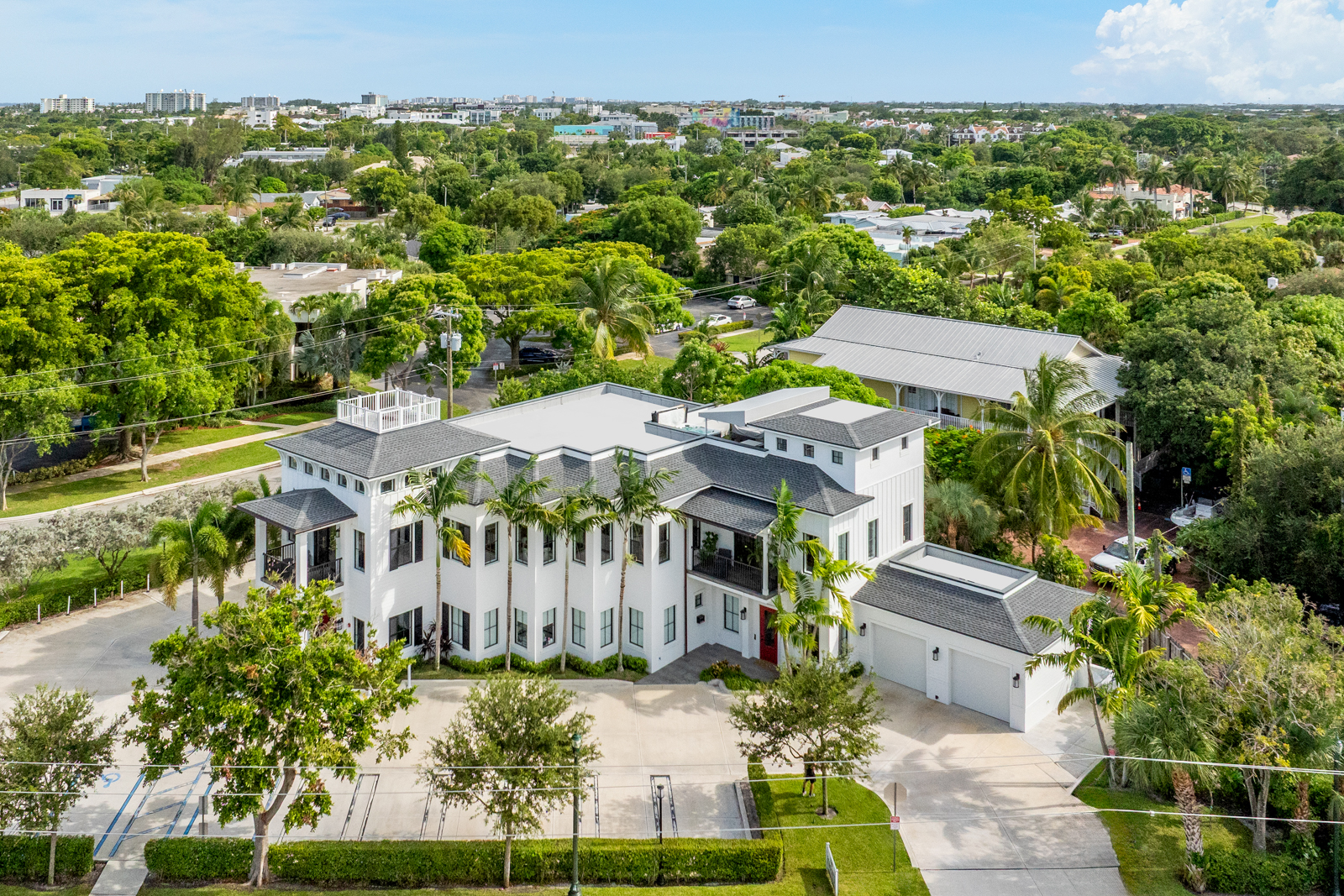 This aerial shot showcases a stunning white modern home with a gray roof, surrounded by lush greenery and palm trees. The property features a spacious driveway, a well-manicured lawn, and a detached garage, creating an inviting and luxurious curb appeal. The overall impression is one of elegance and tranquility, perfect for discerning buyers.