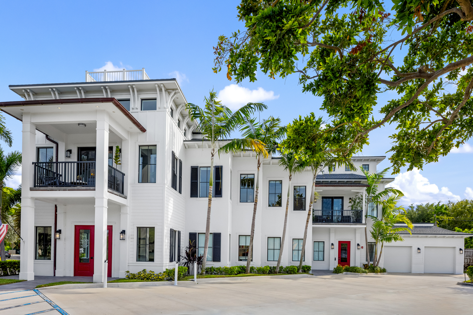 This is a front exterior view of a modern, multi-story building with a white facade, dark-framed windows, and red doors. The building features a balcony, palm trees, and a well-maintained lawn, creating a sophisticated and inviting curb appeal. A two-car garage is attached to the right side of the building.