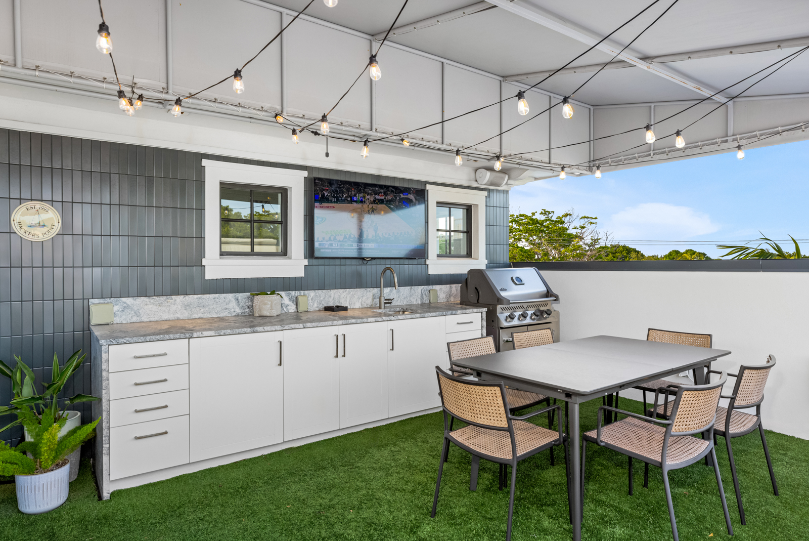 This is a well-appointed outdoor patio area, featuring a built-in kitchen with white cabinetry, a stainless steel grill, and a marble countertop. A dining table with six chairs sits on artificial turf, and string lights hang overhead, creating a cozy atmosphere. A television is mounted on the wall, and the space is partially covered by a roof structure.