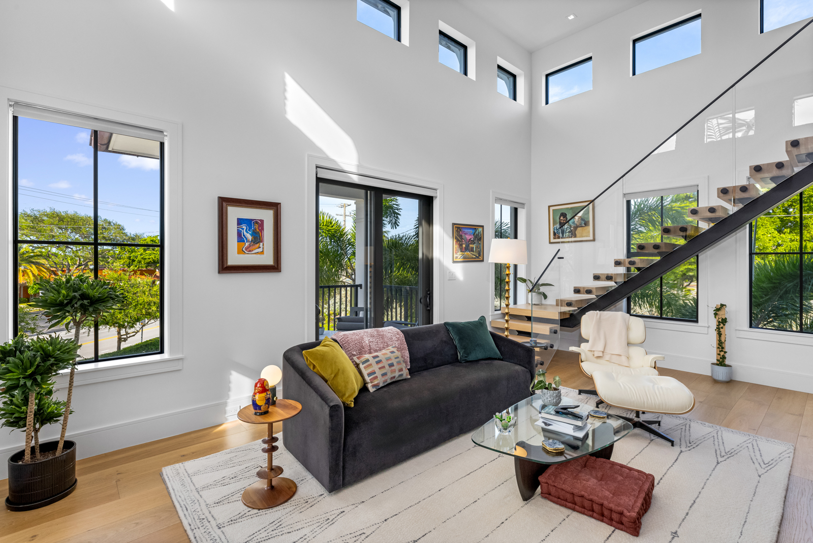 This is an interior shot of a modern living room featuring a dark gray sofa with colorful pillows, a glass coffee table, and a white armchair. The room has a high ceiling with clerestory windows, a staircase with glass railings, and large windows that provide ample natural light. The overall impression is stylish and inviting.