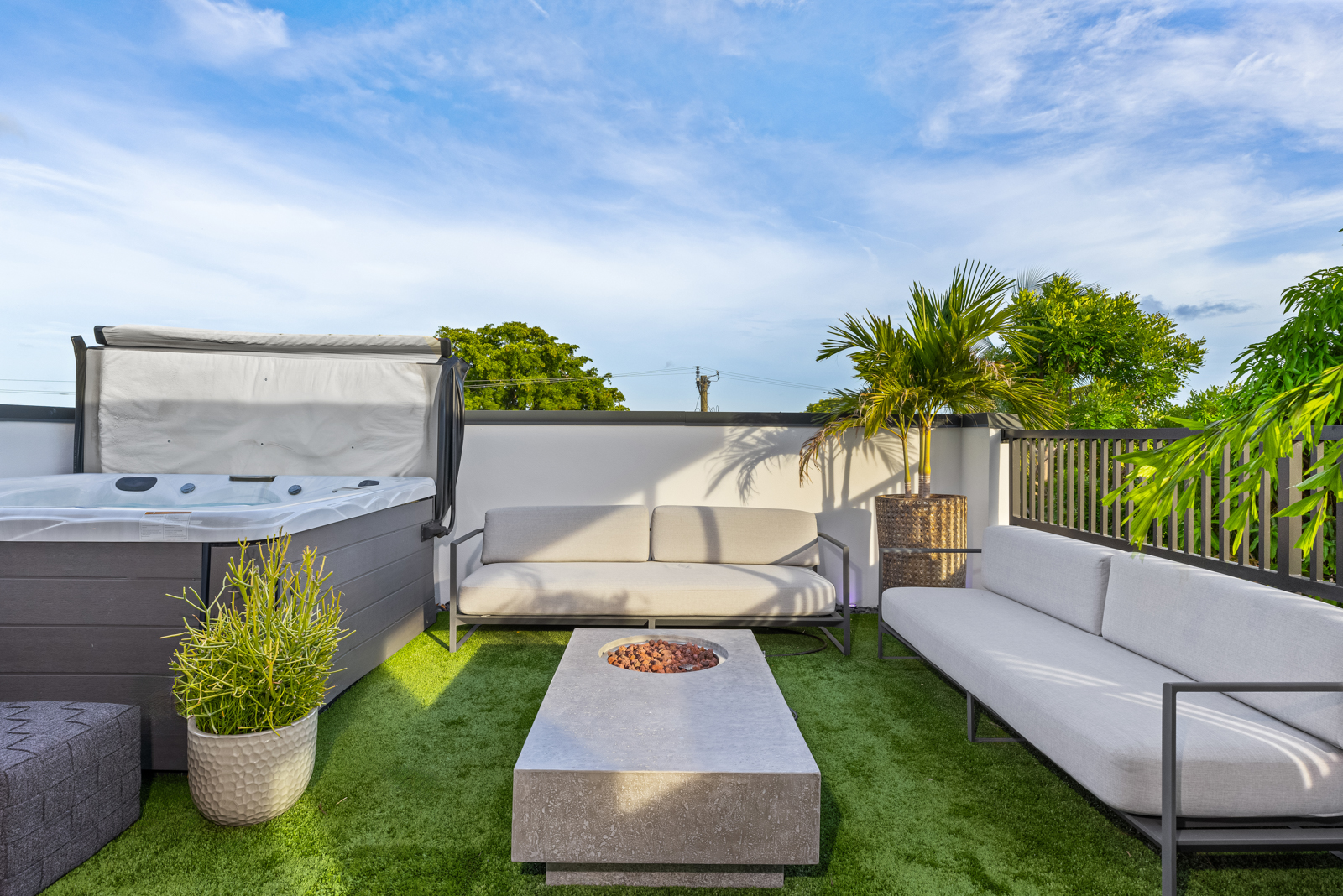 This image showcases a modern rooftop patio or deck, featuring artificial turf, a hot tub, and comfortable outdoor seating. A contemporary fire pit table serves as a focal point, complemented by stylish sofas and potted plants. The space is designed for relaxation and entertainment, offering a luxurious outdoor living experience.