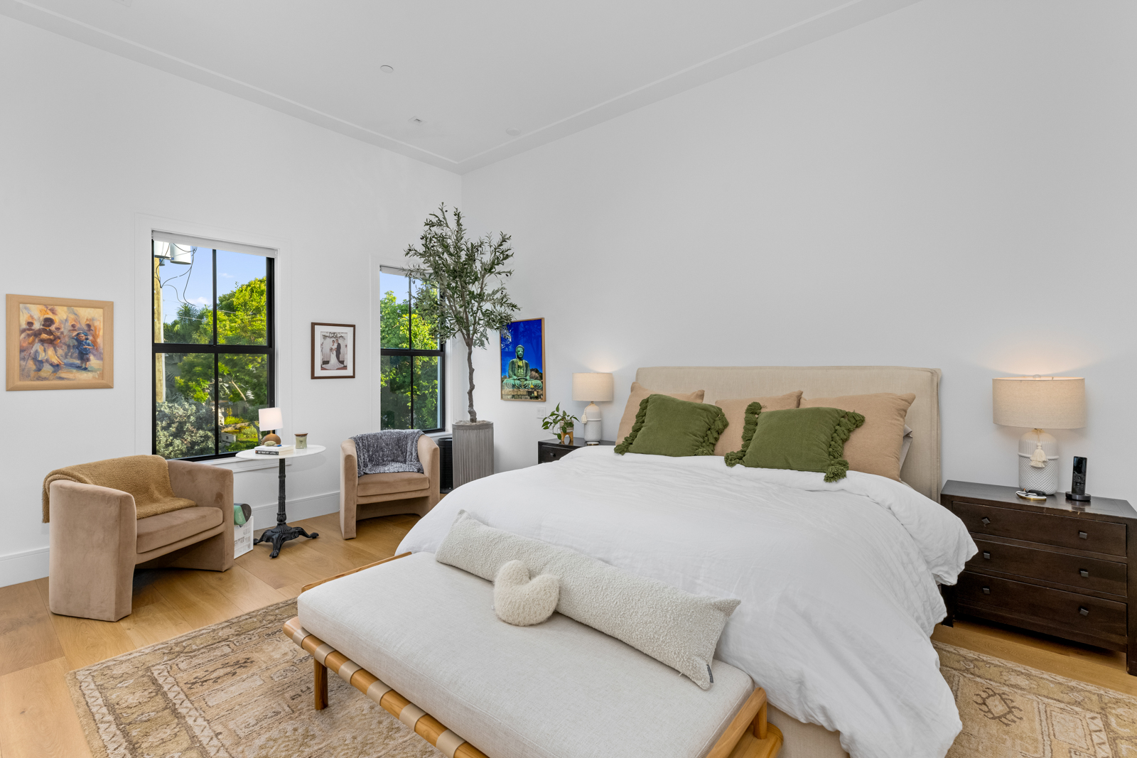 This is a bright and airy primary bedroom featuring a neutral color palette. The room includes a large bed with decorative pillows, two armchairs, and a dark wood nightstand. Natural light floods the space through two windows, enhancing the room's serene and inviting atmosphere. The perspective is a wide shot, capturing the entire room.