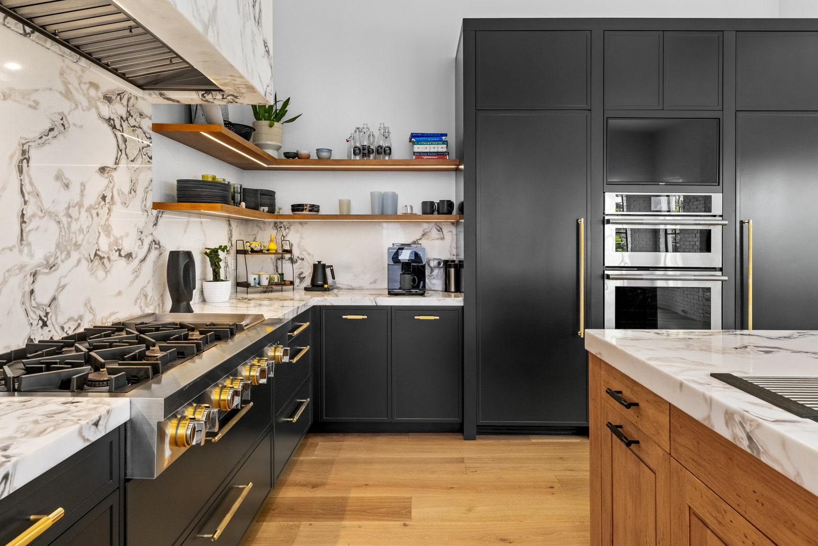 This is a modern kitchen with black cabinetry, gold hardware, and marble countertops and backsplash. The kitchen features a professional-grade range, open shelving, and built-in appliances. The overall impression is luxurious and sophisticated, with a focus on high-end finishes and functionality.