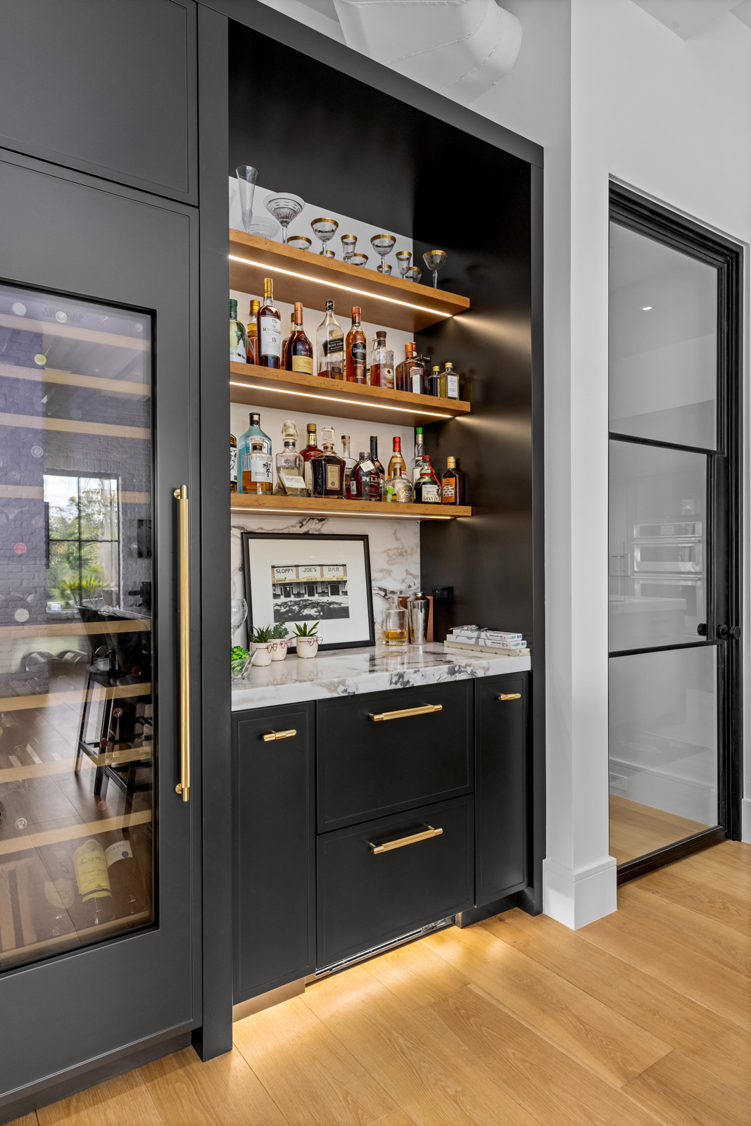 This image showcases a stylish built-in bar area within a modern kitchen. The bar features dark cabinetry with gold hardware, marble countertops, and open shelving displaying glassware and liquor bottles. A wine refrigerator is integrated into the design, adding a touch of luxury.
