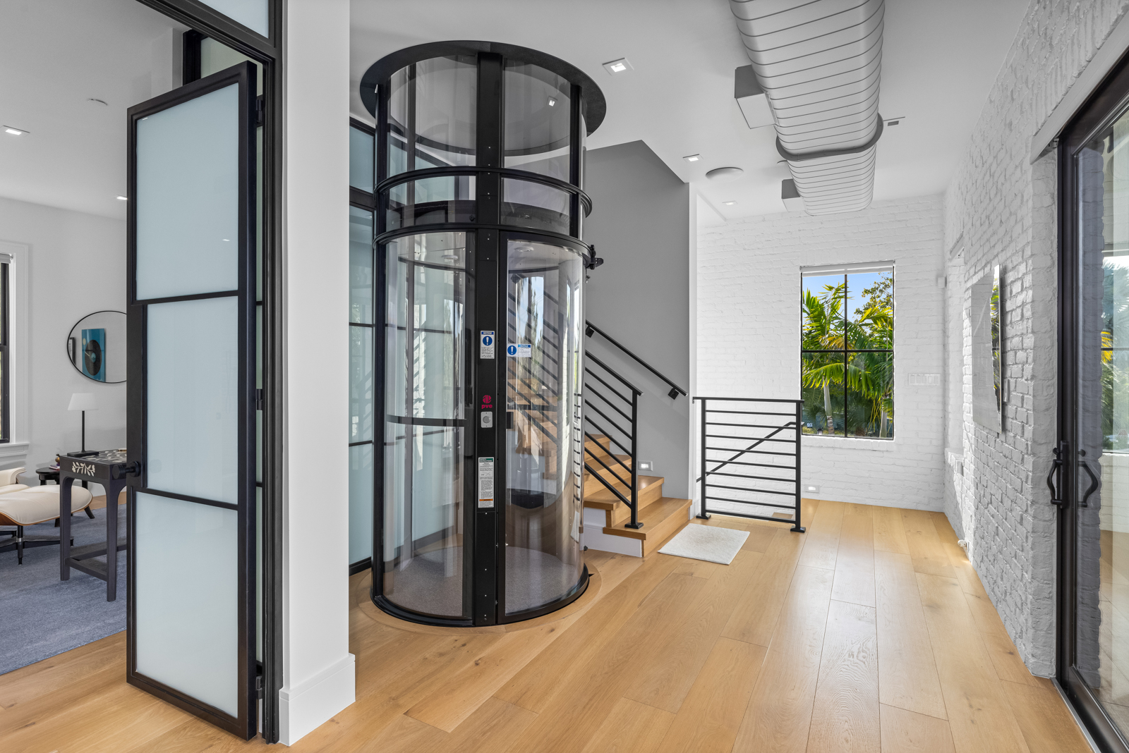 This interior shot showcases a modern hallway featuring light wood flooring and a unique cylindrical glass elevator. A staircase with black railings is visible in the background, leading to an upper level. The hallway is well-lit, with a window providing natural light and a clean, contemporary aesthetic.