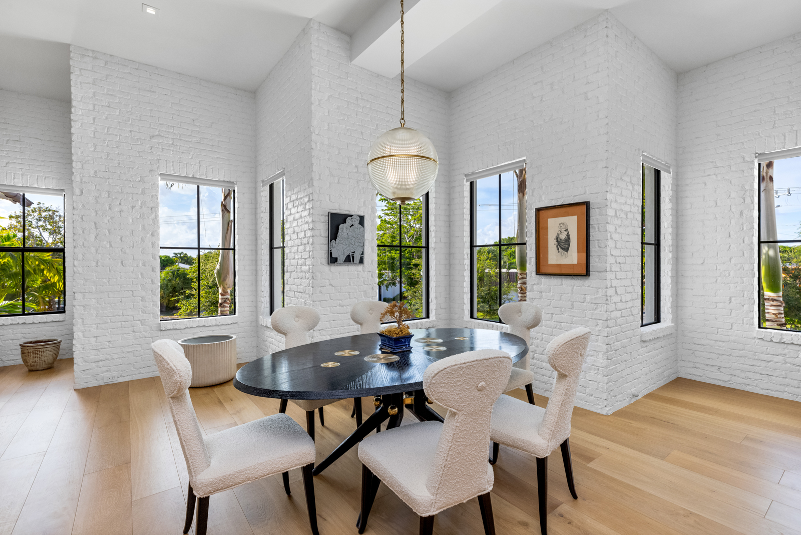 This is an interior shot of a dining room featuring white-painted brick walls and light wood flooring. An oval, dark-colored dining table is surrounded by six light-colored chairs with dark legs. A modern globe pendant light hangs above the table, and several windows with black frames provide natural light and views of the outdoors.
