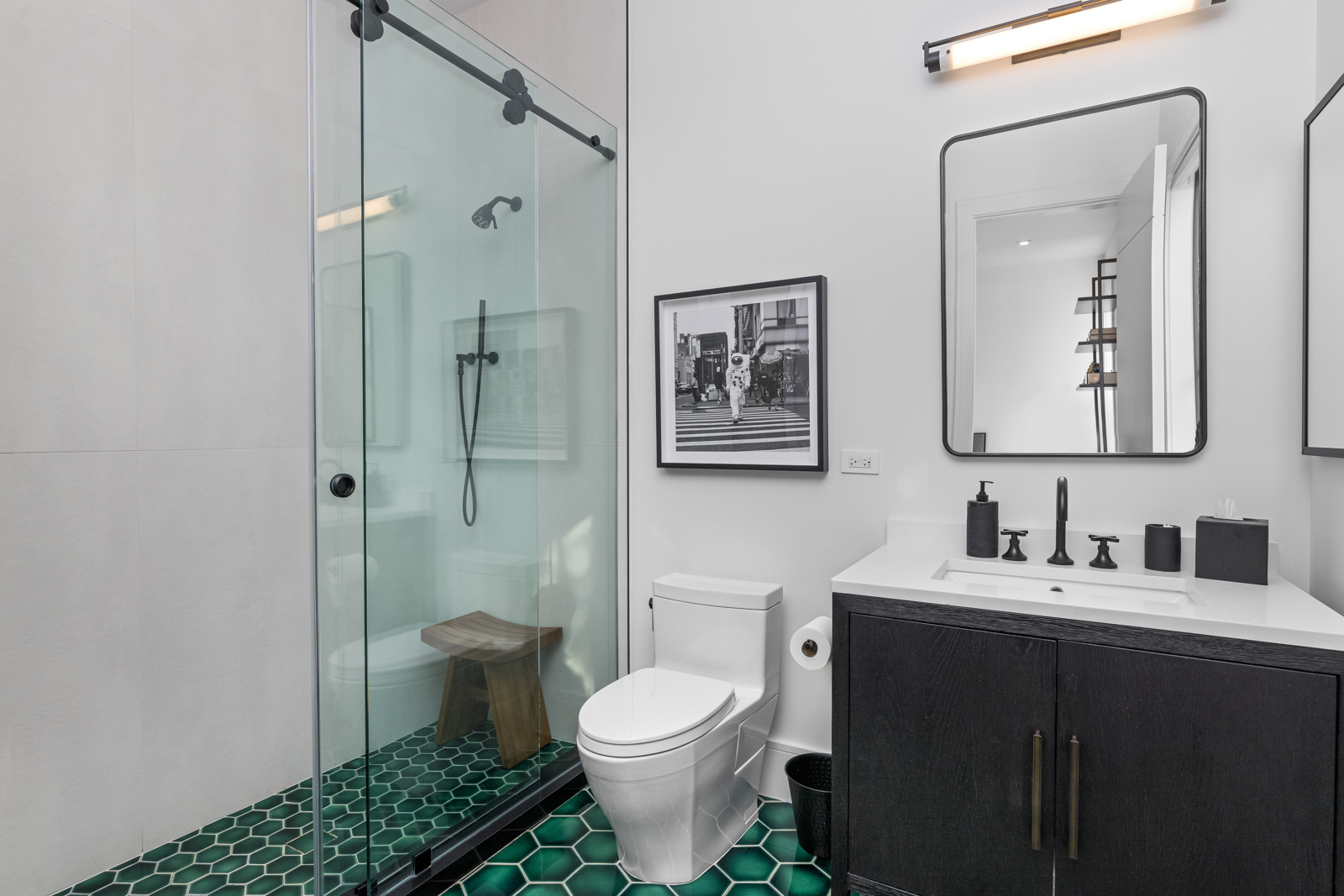 This is a modern guest bathroom featuring a glass-enclosed shower with a black showerhead and a wooden stool inside. The floor is covered in a striking green hexagonal tile, and a white toilet sits next to a black vanity with a white countertop and black fixtures. A framed black and white photograph hangs on the wall, and a rectangular mirror with a light fixture above it completes the contemporary design.