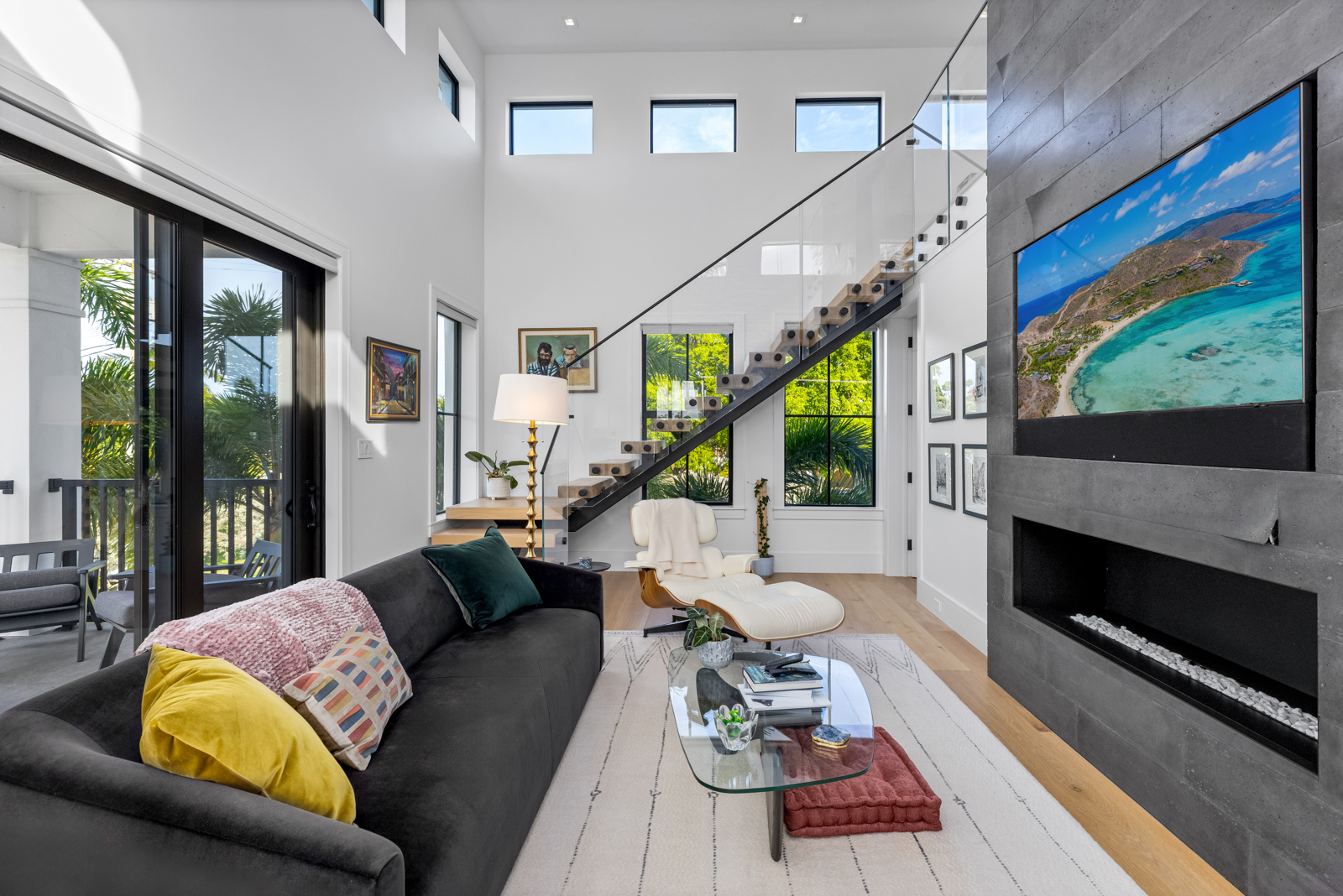 This is a bright and modern living room featuring a dark gray sofa with colorful pillows, a glass coffee table, and a white area rug. A striking staircase with glass railings leads to the upper level, and a contemporary fireplace with a large television above it serves as a focal point. Large windows provide ample natural light, enhancing the airy and spacious feel of the room.
