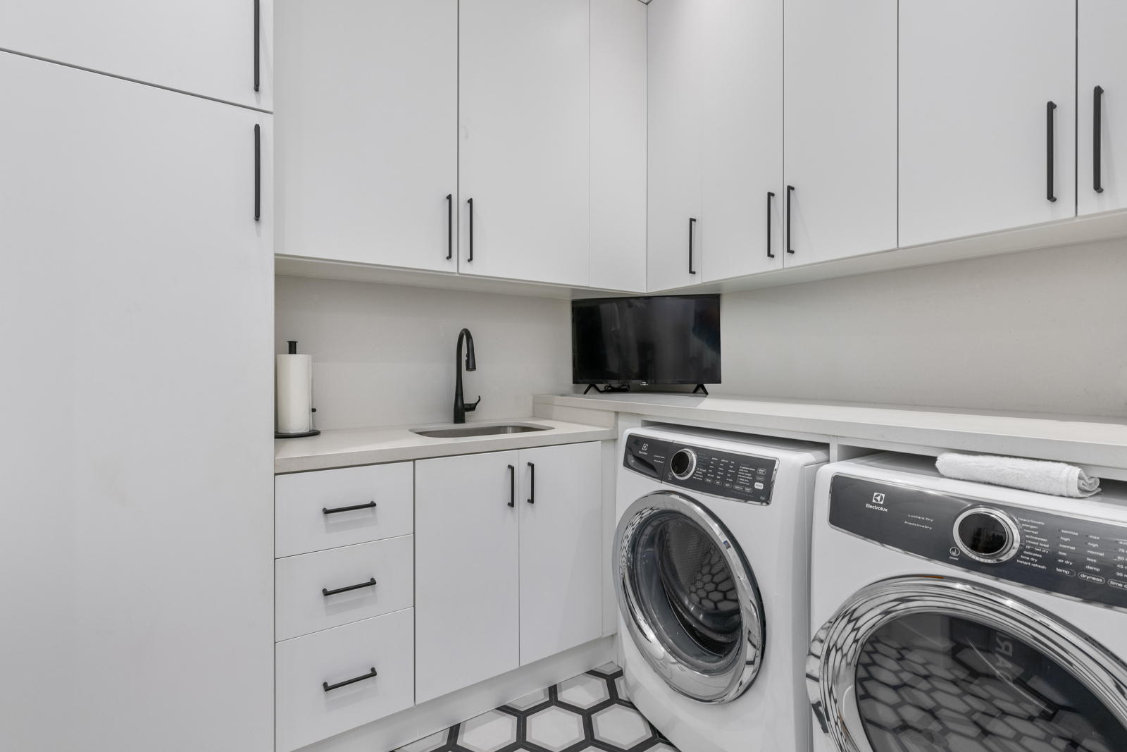 This is a well-organized laundry room featuring white cabinetry with sleek, black hardware and a countertop that extends over the front-loading washer and dryer. A black faucet and stainless steel sink are installed next to a small television. The floor is tiled with a black and white geometric pattern, adding a modern touch to the space.