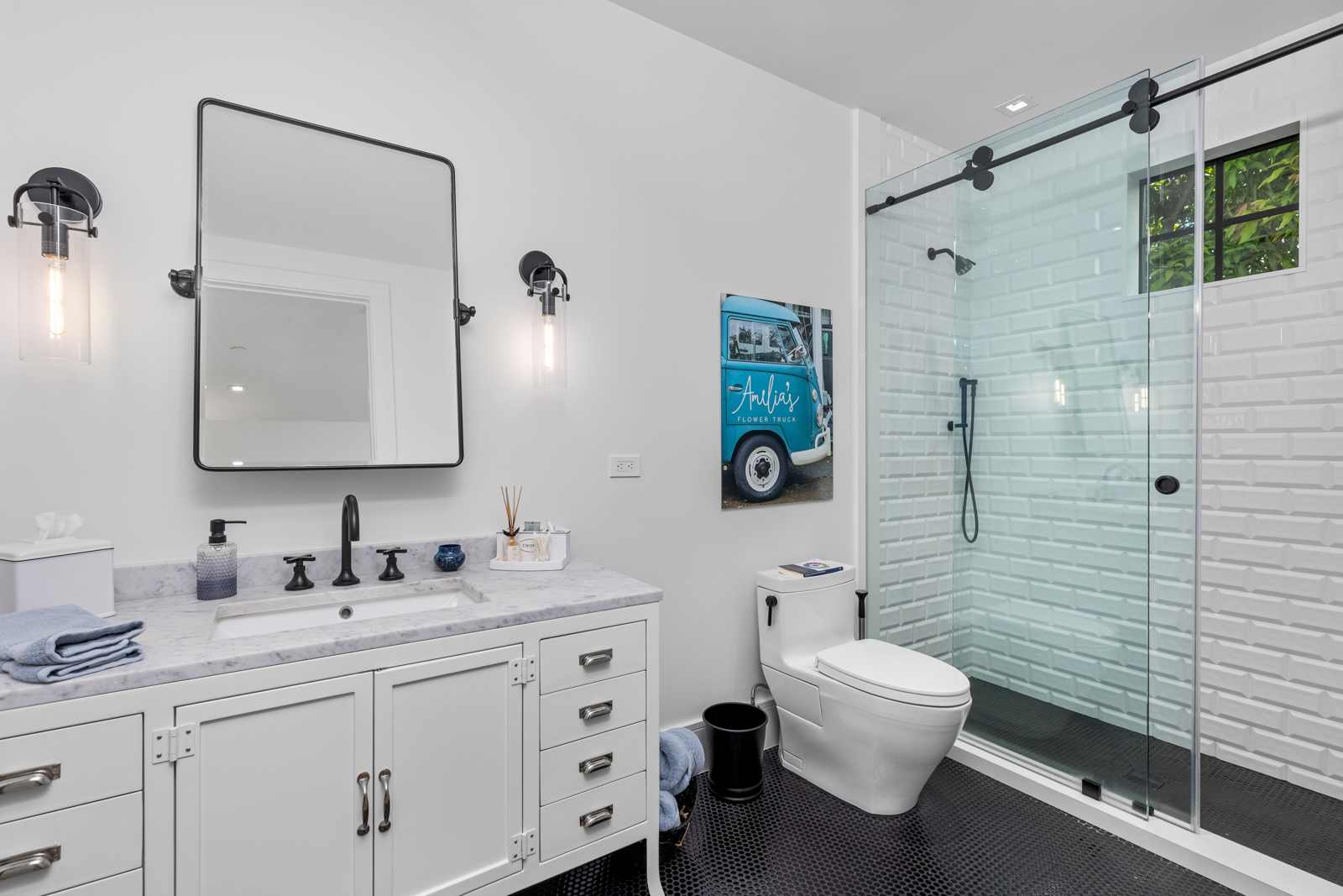 This is a well-lit guest bathroom featuring a white vanity with a marble countertop, a black faucet, and a rectangular mirror with black framing. The shower is enclosed with glass and black hardware, and the walls are tiled with white subway tiles. The floor is covered in black penny tiles, creating a modern and clean aesthetic.