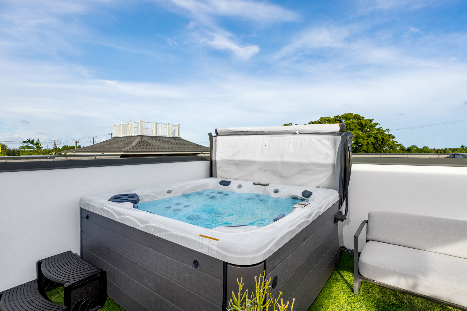 This image showcases a modern spa on a rooftop deck, featuring a sleek gray and white hot tub filled with clear blue water. The surrounding area includes artificial turf, a small seating area, and a partial view of the neighborhood, creating a private and luxurious outdoor space. The open sky and clean design enhance the appeal of this relaxing amenity.
