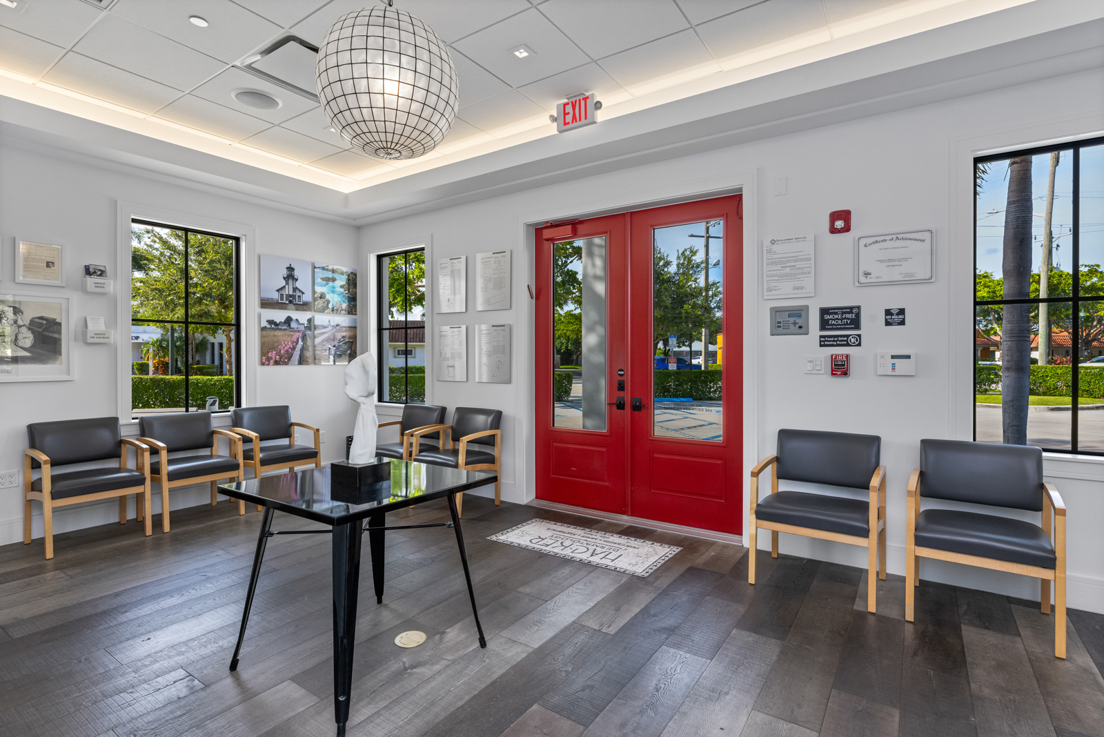 This is an interior shot of an office waiting room featuring a modern design with gray hardwood floors and white walls. The room is furnished with gray chairs with wooden frames, a black table with a white sculpture, and a red double door. Natural light streams in through the windows, creating a bright and inviting atmosphere.