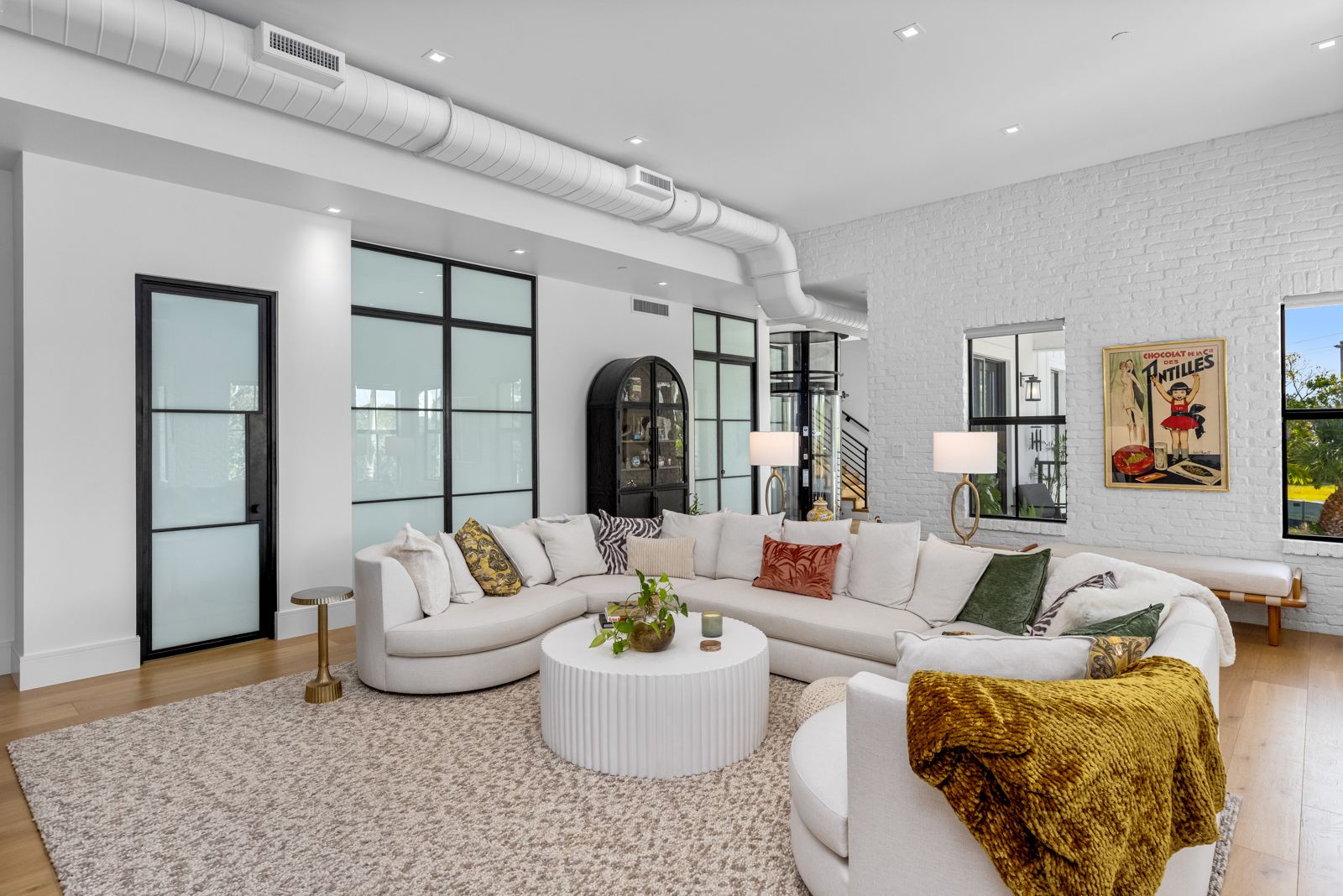 This is a bright and airy living room featuring a large, curved white sofa arranged around a round, fluted coffee table. The room is decorated with a mix of textures and patterns, including throw pillows and a cozy throw blanket. Large windows and black-framed glass doors provide ample natural light, highlighting the hardwood floors and neutral-toned rug.