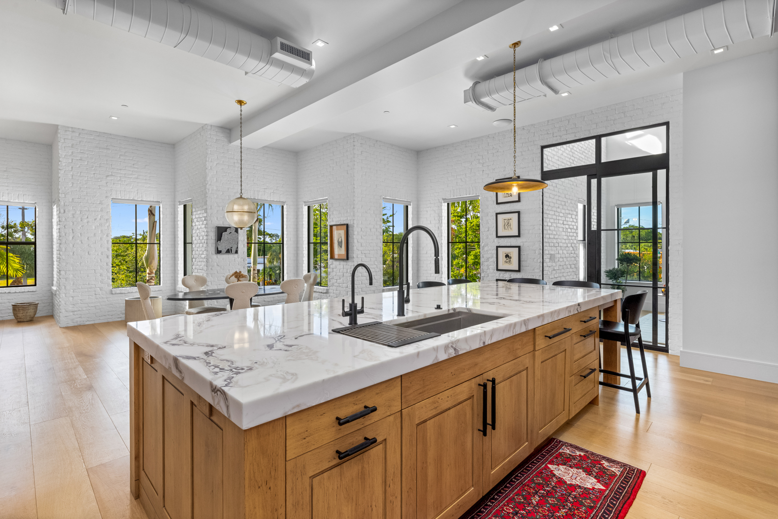 This is a bright and modern kitchen featuring white-painted brick walls and light wood flooring. A large kitchen island with a marble countertop and wood cabinetry dominates the space, equipped with a sink and black fixtures. The room is well-lit with natural light from several windows and pendant lighting, creating an inviting and stylish atmosphere.