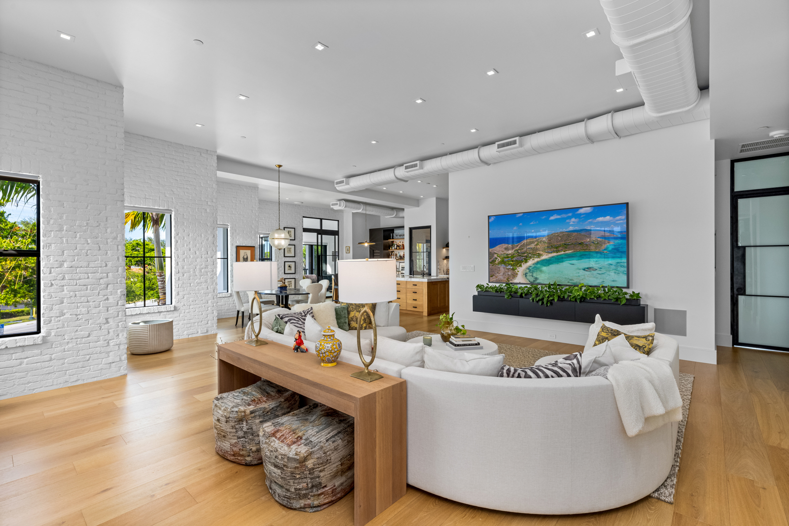 This is a bright and airy living room featuring a large, curved white sofa as the focal point. The room has hardwood floors, white brick accents, and large windows that provide ample natural light. A console table sits behind the sofa, adorned with lamps and decorative items, while a large television is mounted on the wall, complemented by greenery below.