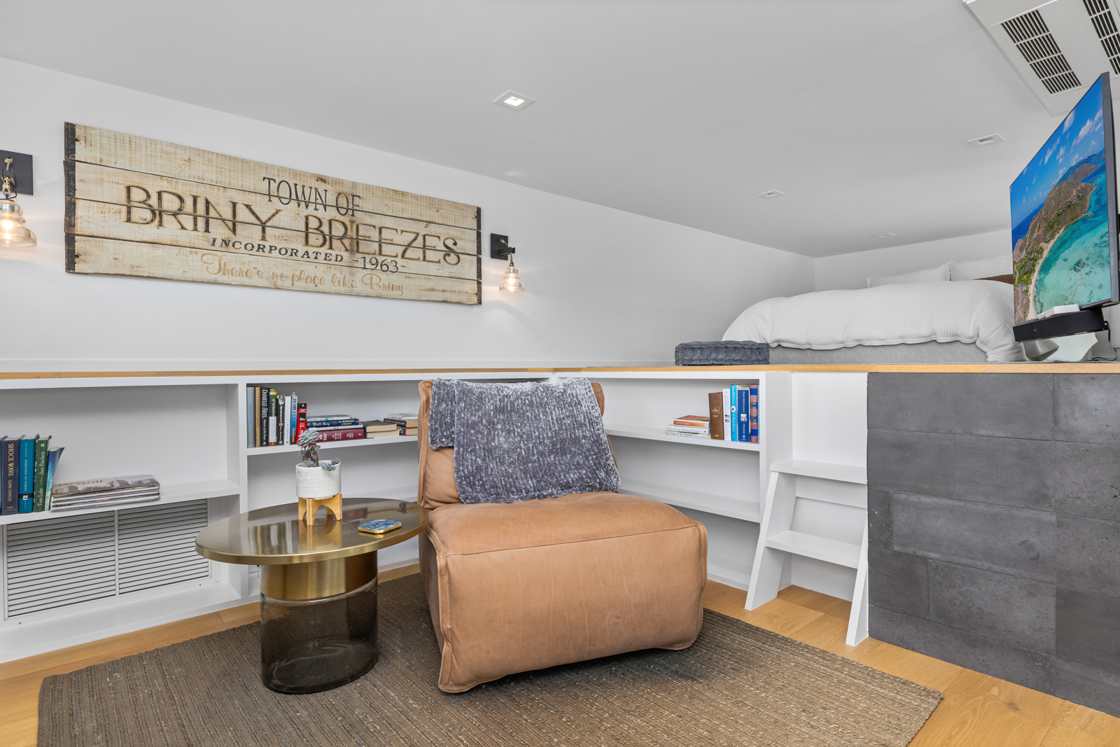 This interior shot showcases a cozy bedroom nook featuring a comfortable armchair with a throw blanket, a round side table, and built-in shelving filled with books. Above, a lofted bed area is visible, complete with a television. The space is well-lit and designed for relaxation, creating an inviting atmosphere.