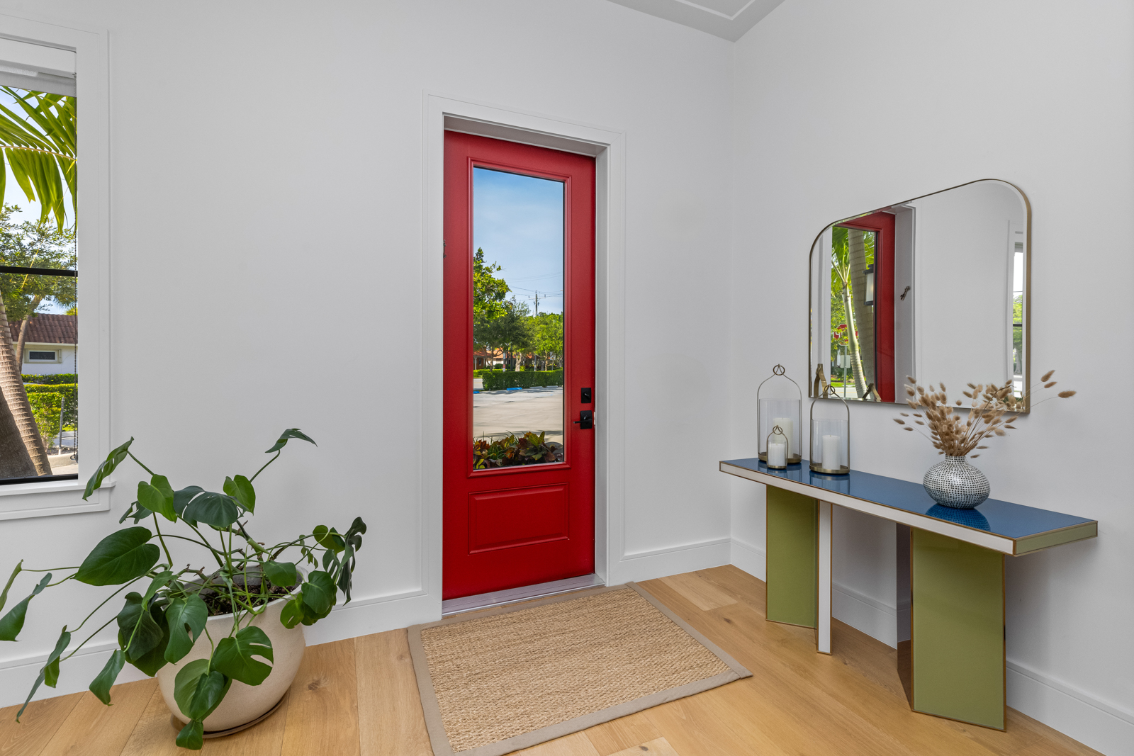 This interior shot showcases a bright and welcoming entryway with a vibrant red door, complemented by a neutral-toned rug and light wood flooring. A modern console table with a mirror and decorative items adds a touch of elegance, while a potted plant near the window brings a natural element to the space. The walls are painted in a clean white, enhancing the sense of spaciousness and light.