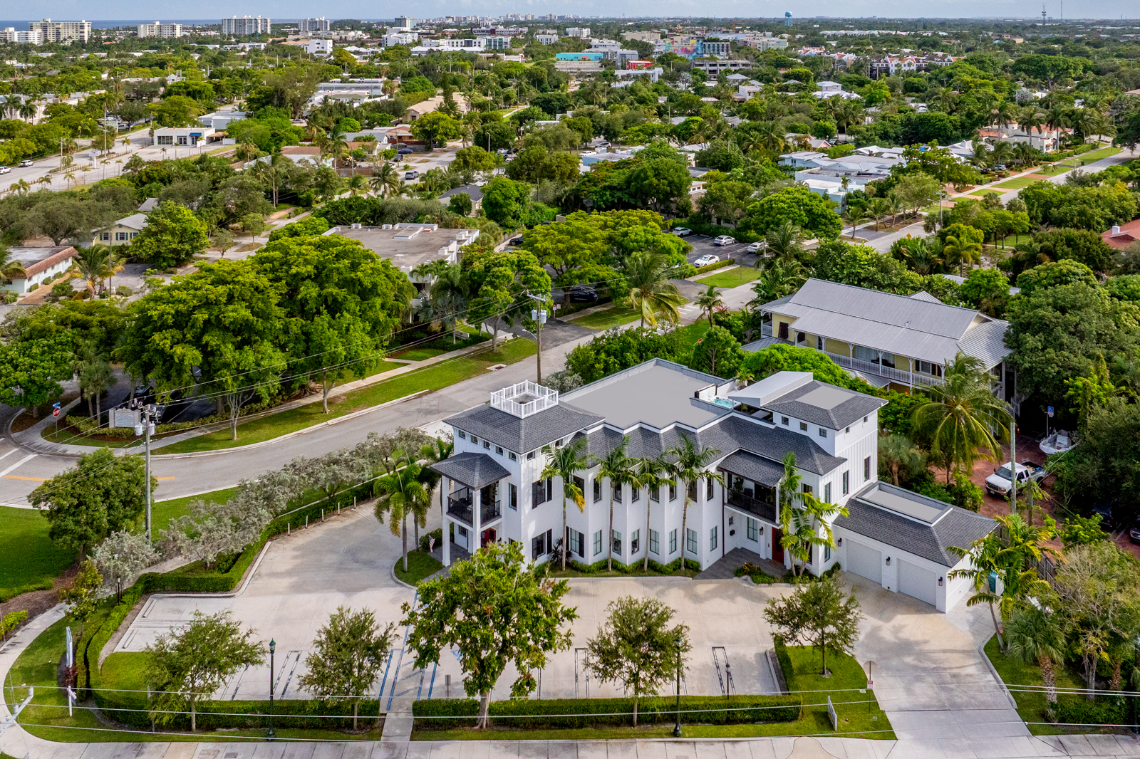 This aerial shot showcases a grand, two-story white building with a dark gray roof, surrounded by lush greenery and palm trees. A spacious parking area is visible in front, and the building's architectural details, including balconies and a rooftop terrace, are prominently displayed. The image provides a comprehensive view of the property and its setting within a residential neighborhood.